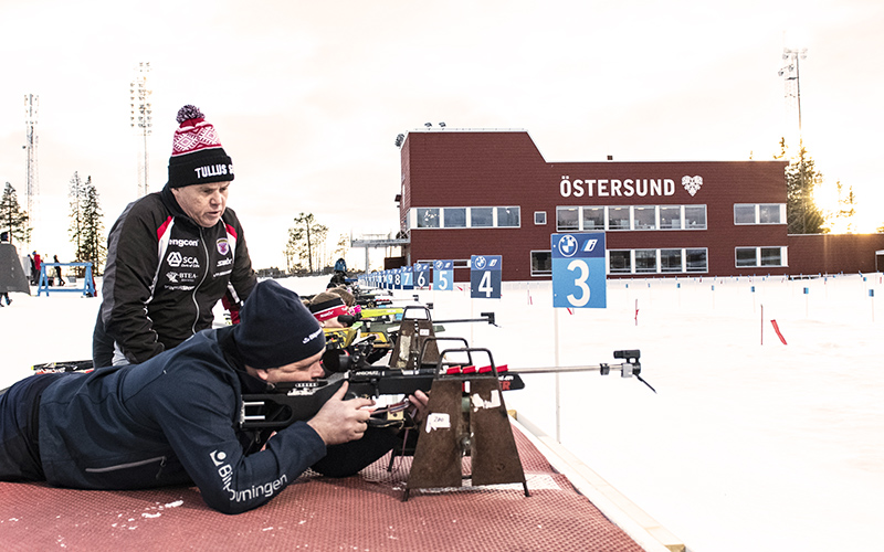 En man iklädd bilprovningskläder ligger och skjuter på skidstadion i Östersund. Bredvid syns en instruerande man och i bakgrund ungdomar som också ligger och skjuter. 