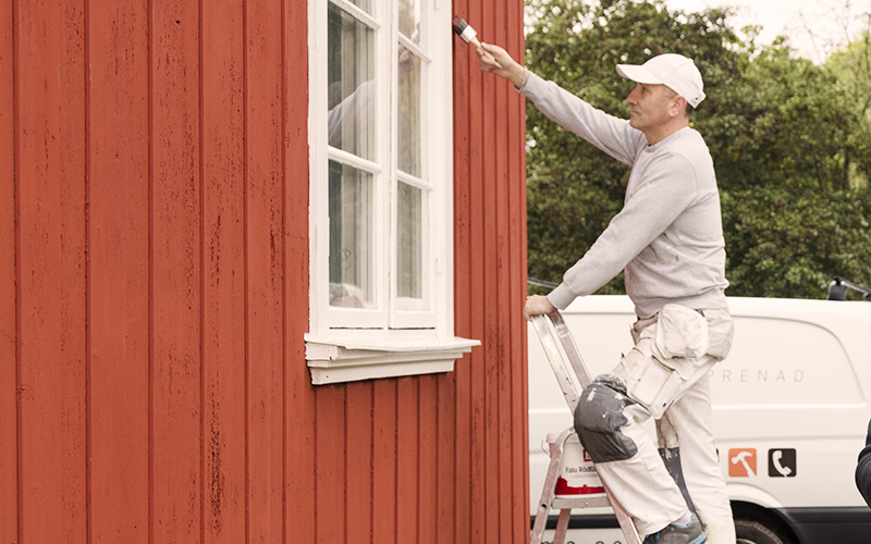 En hantverkare står på en stege och målar en fasad. I bakgrunden syns en vit lätt lastbil. 
