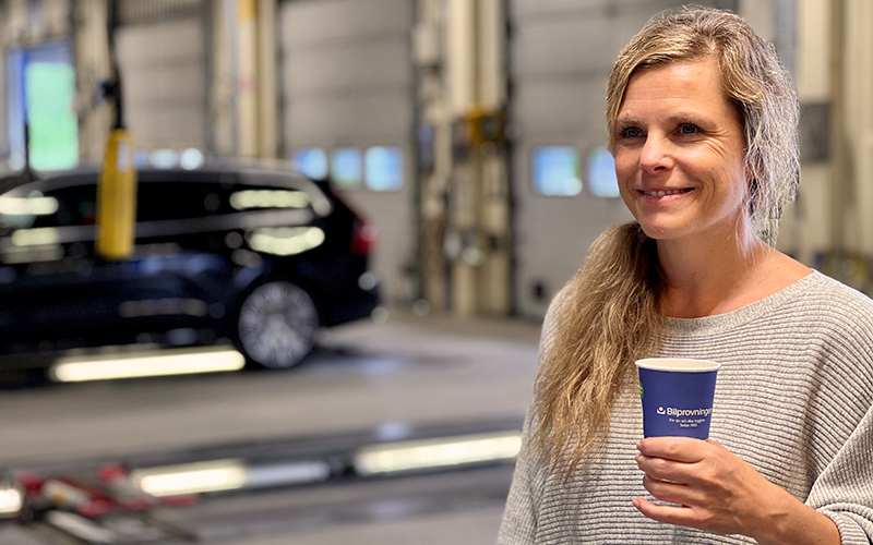 En kvinna står med en kopp märkt med Bilprovningens logotyp i en besiktningshall framför lyftarna. 