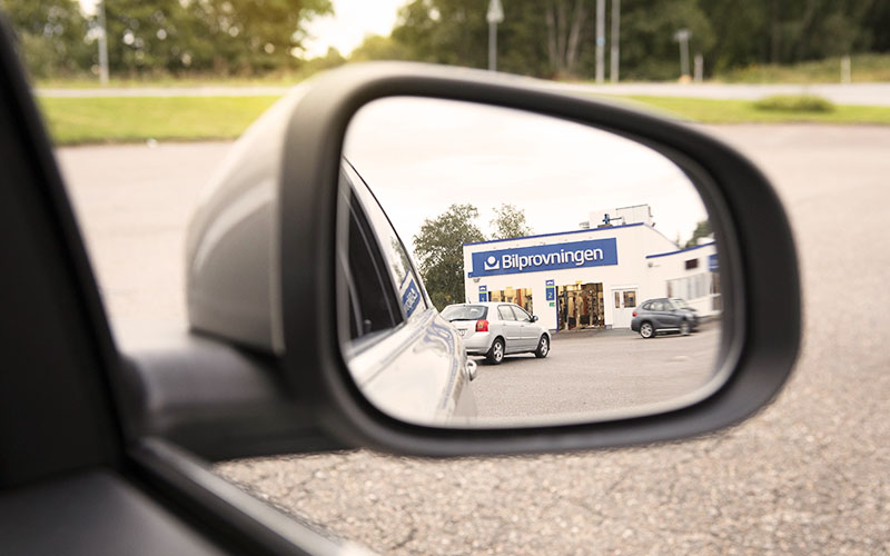 En bilprovningsstations exteriör och parkering med personbilar syns i en sidospegel på en personbil.