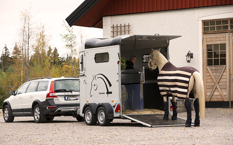 Kvinna leder en häst in i en hästtransport som dras efter en vit bil och står parkerad utanför ett stall