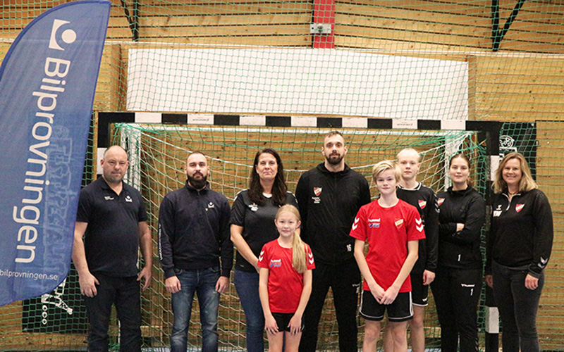 Medarbetare från Bilprovningen står tillsammans med vuxna och barn från handbollsföreningen Skånela IF i idrottshallen. 