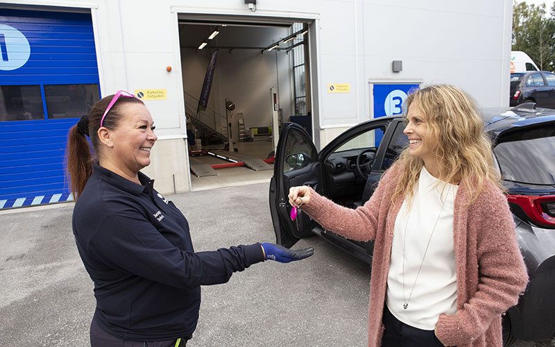 En kvinna i bilprovningskläder tar emot nycklar av en annan kvinna framför en bilprovningsanläggning