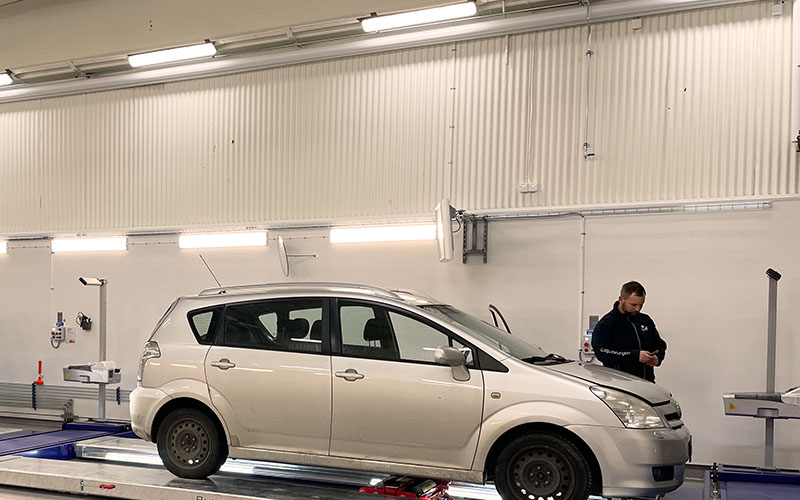 En manlig tekniker besiktar en vit personbil i en LED-upplyst besiktningshall. 